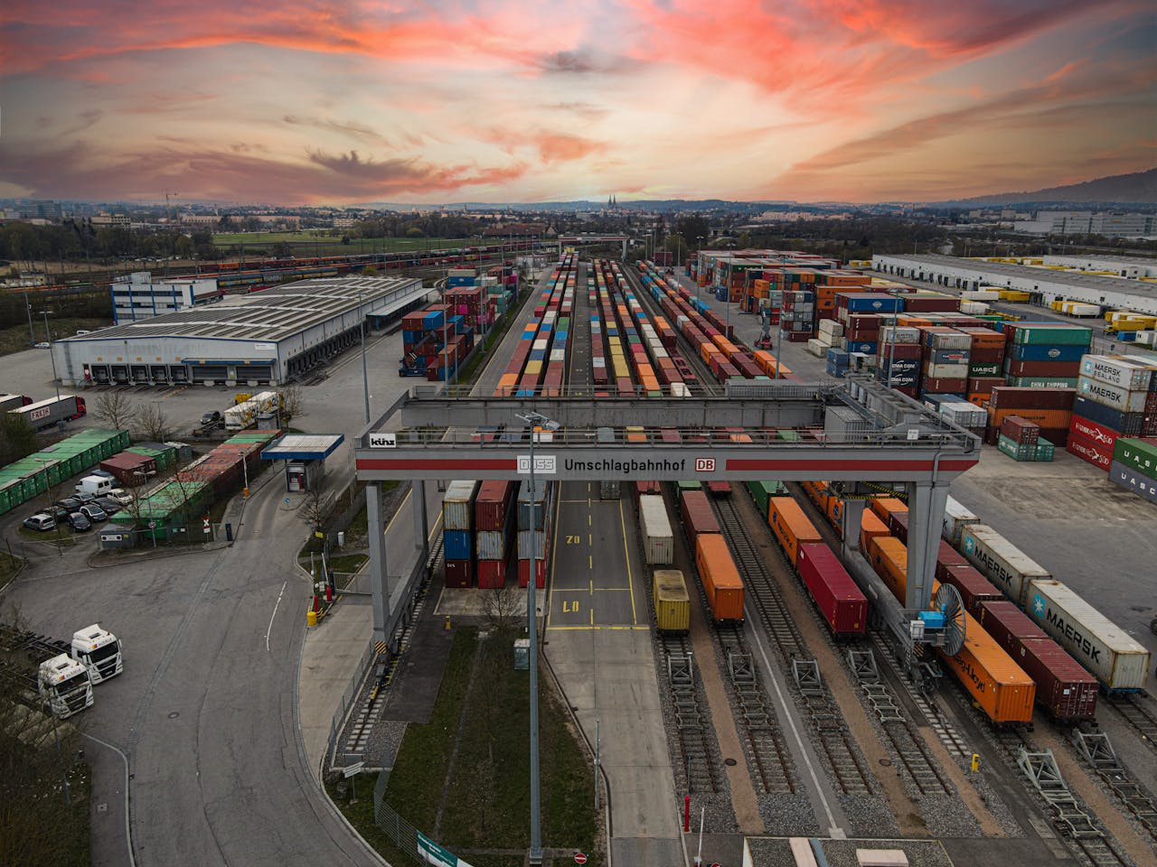 hero-img-02 Aerial view of a bustling shipping yard with colorful containers at sunset, highlighting logistics.